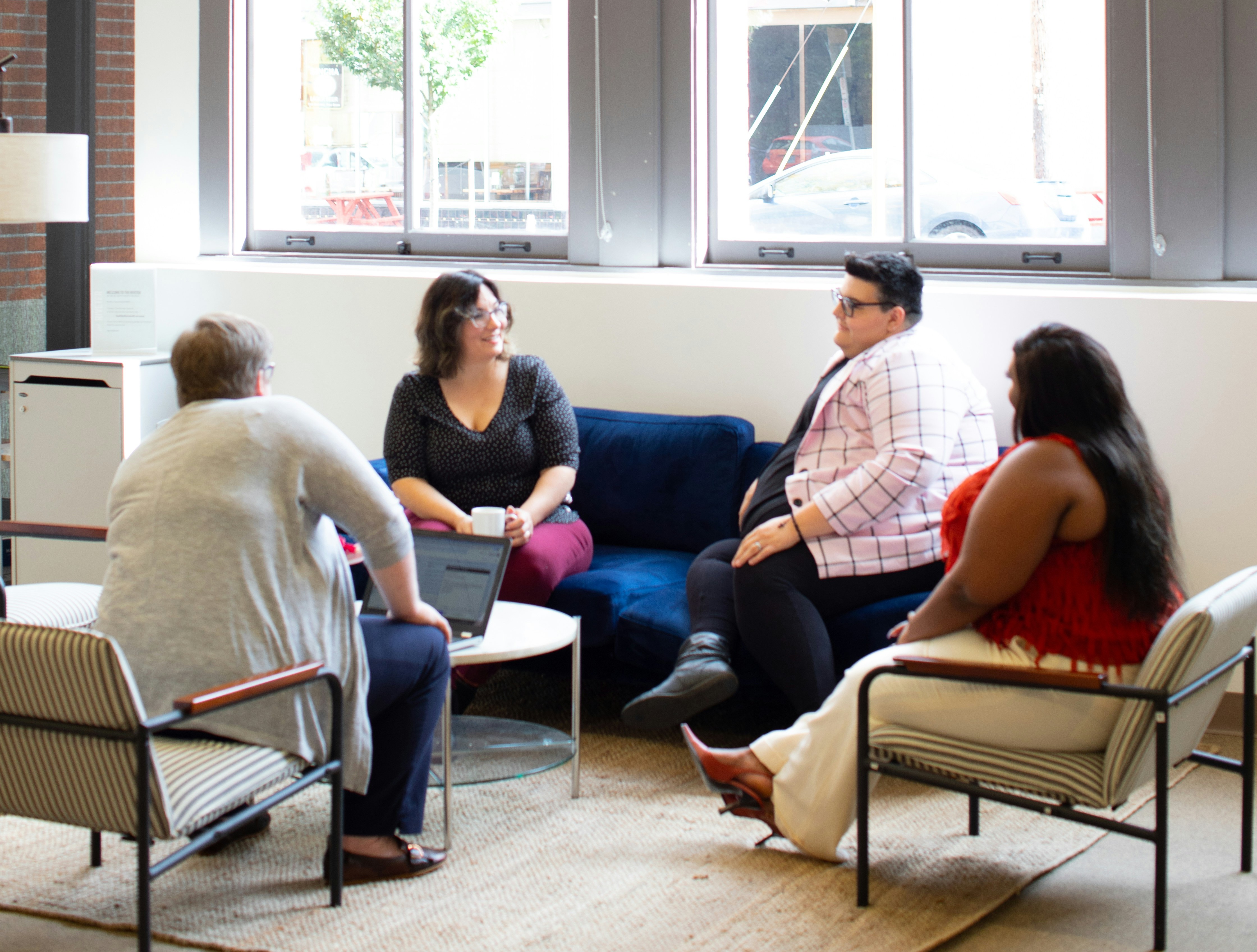 a group of four women sit in a bright living room with windows. They have coffee and are speaking to each other.